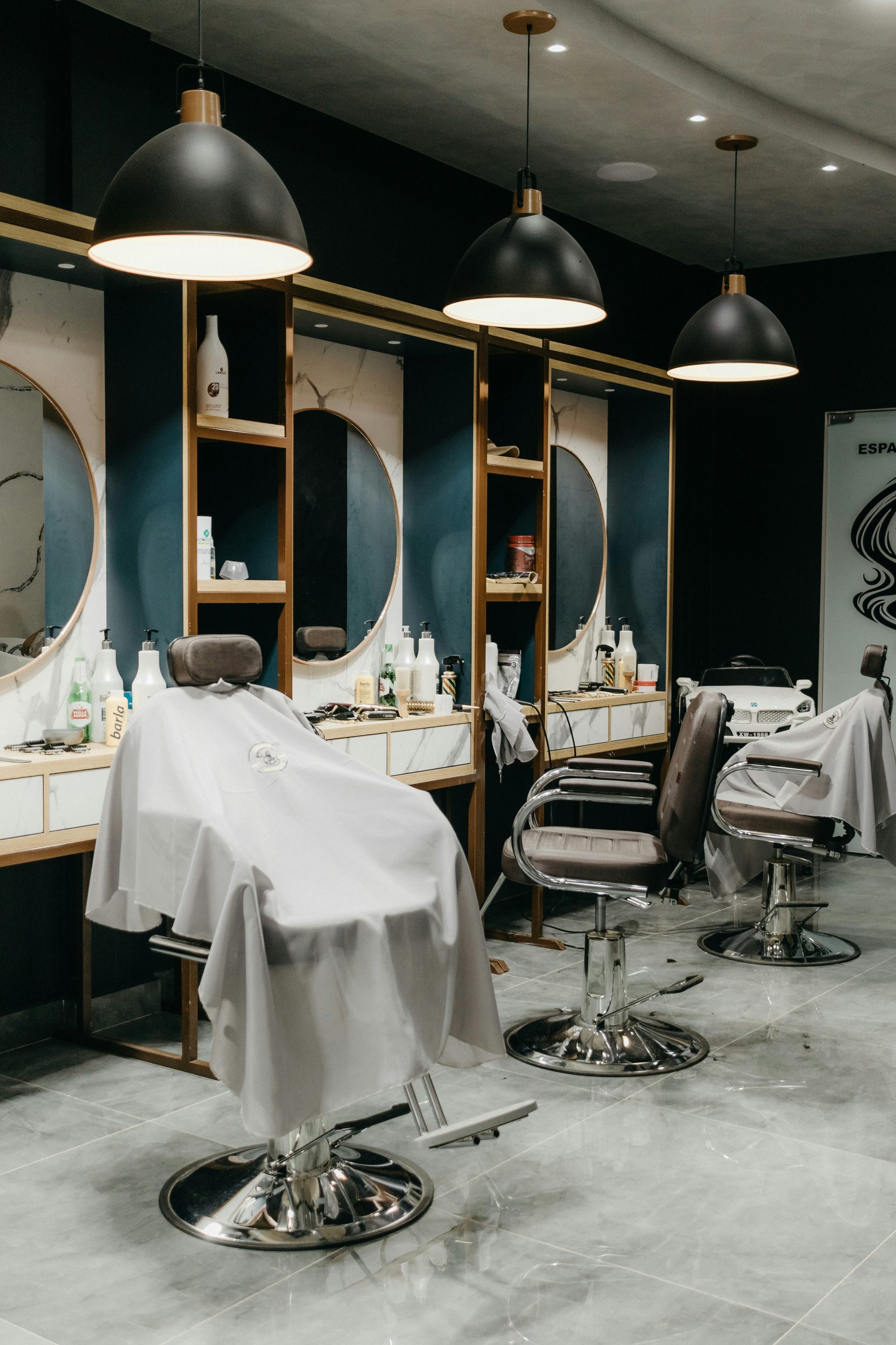 Modern barbershop with three empty chairs draped in white capes. Large round mirrors, wooden shelving, and pendant lights create a sleek ambiance.